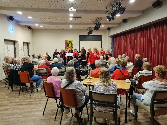 Foto van een groep mensen bij elkaar in een gemeenschapshuis, luisterend naar een koor.